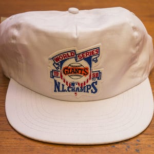 Pu&ograve; includere: Cappellino da baseball bianco con una toppa blu, rossa e bianca che dice "World Series '89 Giants '89 N.L. Champs".