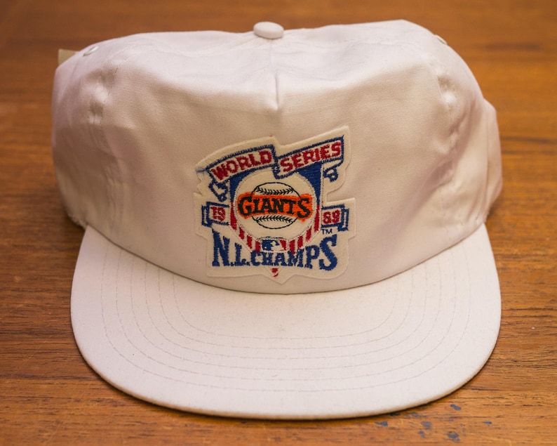 Pu&ograve; includere: Cappellino da baseball bianco con una toppa blu, rossa e bianca che dice "World Series '89 Giants '89 N.L. Champs".
