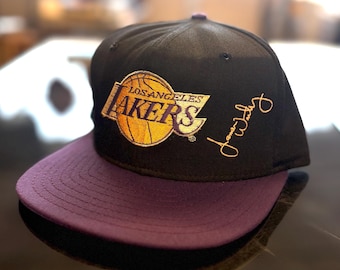 May include: A black baseball cap with a purple flat brim featuring the embroidered Los Angeles Lakers logo in yellow and purple on the front crown. The cap includes a gold embroidered signature on the side panel near the team logo.