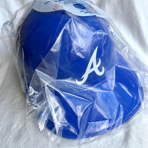Op de afbeelding: Een blauwe baseballcap met een wit "A" logo, verpakt in doorzichtig plastic. De cap heeft een gebogen klep. De verpakking heeft gedeeltelijk zichtbare tekst. Het is een sportartikel.