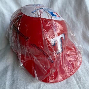 Op de afbeelding: Rode honkbalpet met een wit en blauw "T" logo, verpakt in een doorzichtige plastic zak. De bovenkant van de pet heeft een wit gedeelte met tekst en een honkbalafbeelding.