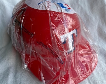 May include: A red baseball cap with the letter "T" in white and blue on the front. The cap is in a clear plastic bag. The top of the cap has a white section with text and a baseball design.
