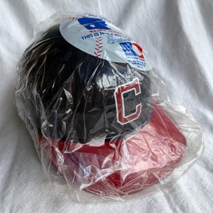 Op de afbeelding: Een zwarte baseball cap met een rode klep en een wit omlijnde "C" op de voorkant. De cap zit in een doorzichtige plastic zak met "Major League Baseball" branding.