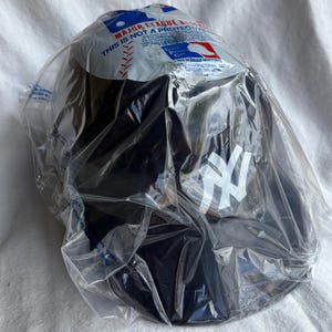 Op de afbeelding: Een zwarte baseballcap met het witte logo van de New York Yankees, verpakt in doorzichtig plastic. De verpakking bevat tekst en een Major League Baseball-logo.