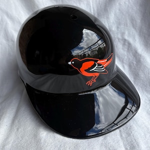 Op de afbeelding: Een glanzende zwarte honkbalhelm met een gebogen vizier. Een rood en oranje vogel logo staat op de zijkant. De helm reflecteert de lucht en de omgeving.