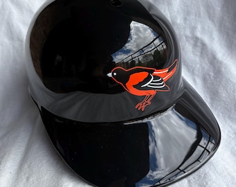 May include: A glossy black baseball helmet with a curved visor. A red and orange bird logo is on the side. The helmet reflects the sky and surrounding environment.