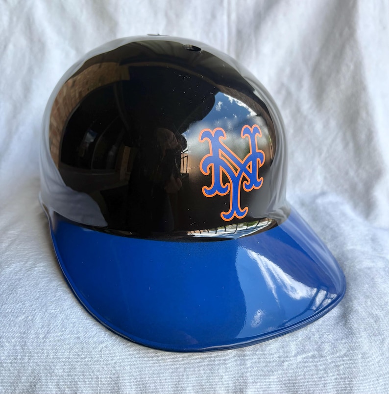 Op de afbeelding: Een glanzende zwarte en blauwe honkbalhelm met het New York Mets-logo in oranje en blauw. De helm heeft een gebogen vizier en is ontworpen voor bescherming tijdens honkbalwedstrijden. De helm is een sportaccessoire.