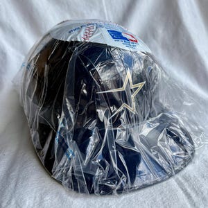 Op de afbeelding: Een marineblauwe baseballpet met een gouden sterlogo, verpakt in doorzichtig plastic. De pet heeft een gebogen klep en is waarschijnlijk nieuw. De verpakking bevat tekst en een honkbalafbeelding.