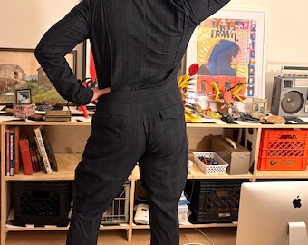 May include: A dark charcoal gray jumpsuit featuring a subtle white windowpane plaid pattern, long sleeves, a collared neckline, and two back pockets. The garment is fitted through the torso and legs, ending at the ankles with black shoes.
