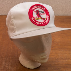 May include: White baseball cap with a red, white and blue patch that says "Cardinals 1985 N.L. East Champs".