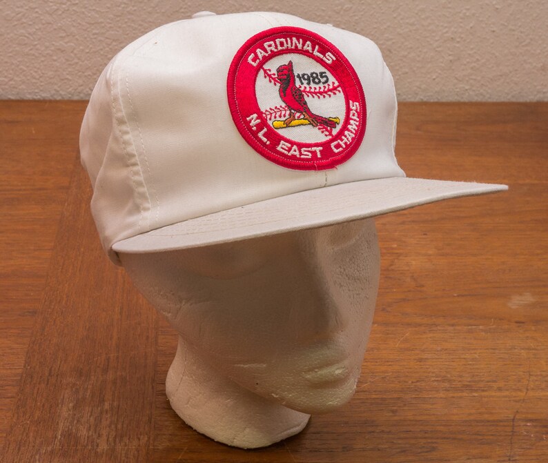 May include: White baseball cap with a red, white and blue patch that says "Cardinals 1985 N.L. East Champs".