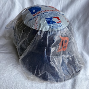 Op de afbeelding: Een marineblauwe honkbalhelm in een doorzichtige plastic zak. De helm heeft een oranje "D"-logo en is gemarkeerd met "Major League Baseball". De verpakking bevat de tekst "Dit is geen beschermende helm."
