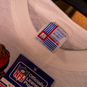 May include: Close-up of a white t-shirt with a tag that reads "OFFICIALLY LICENSED PRODUCT by GARAN, Inc." The tag has red and blue stripes. An NFL tag is visible. The shirt has a graphic of a football.