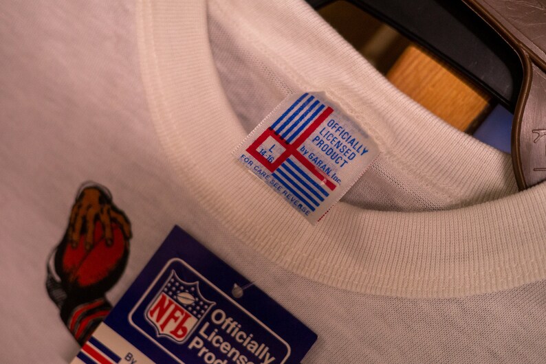May include: Close-up of a white t-shirt with a tag that reads "OFFICIALLY LICENSED PRODUCT by GARAN, Inc." The tag has red and blue stripes. An NFL tag is visible. The shirt has a graphic of a football.