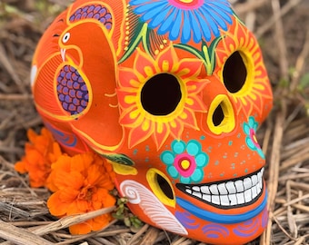 May include: A bright orange ceramic skull sculpture featuring hand-painted floral patterns in blue, yellow, and pink, along with a stylized bird motif. The skull has hollow eye sockets, a nasal cavity, and a wide grin with white teeth, accented by orange flowers.