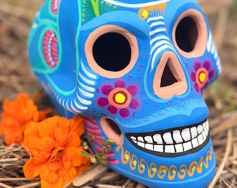 May include: A bright blue ceramic skull sculpture featuring hand-painted floral patterns in purple, yellow, and white. The skull has large eye sockets, a triangular nose cavity, and a wide grin with individual white teeth painted on the jaw.