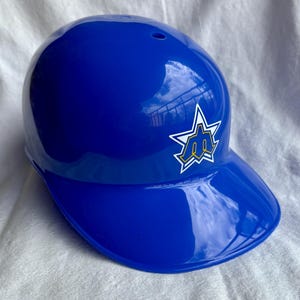 Op de afbeelding: Een glanzende, koningsblauwe honkbalhelm met een vizier. Een wit stervormig logo met een geel en blauw embleem staat op de zijkant. De helm is ontworpen om het hoofd te beschermen tijdens honkbalwedstrijden.