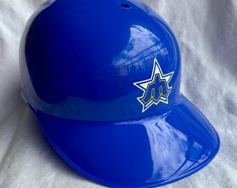 May include: A glossy, royal blue baseball helmet with a visor. A white star-shaped logo with a yellow and blue emblem is on the side. The helmet is designed to protect the head during baseball games.