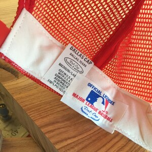 Puede incluir: Gorra de b&eacute;isbol de malla roja con una etiqueta blanca que dice "Dallas Cap, Dallas, Texas, Dal-Cap, Medium/Lar, Cut in U.S.A., Component with U.S.A., Assembled in Mexico, 100% Polyester" y una etiqueta blanca que dice "Official Licensee, Major League Baseball, Dal-Cap".