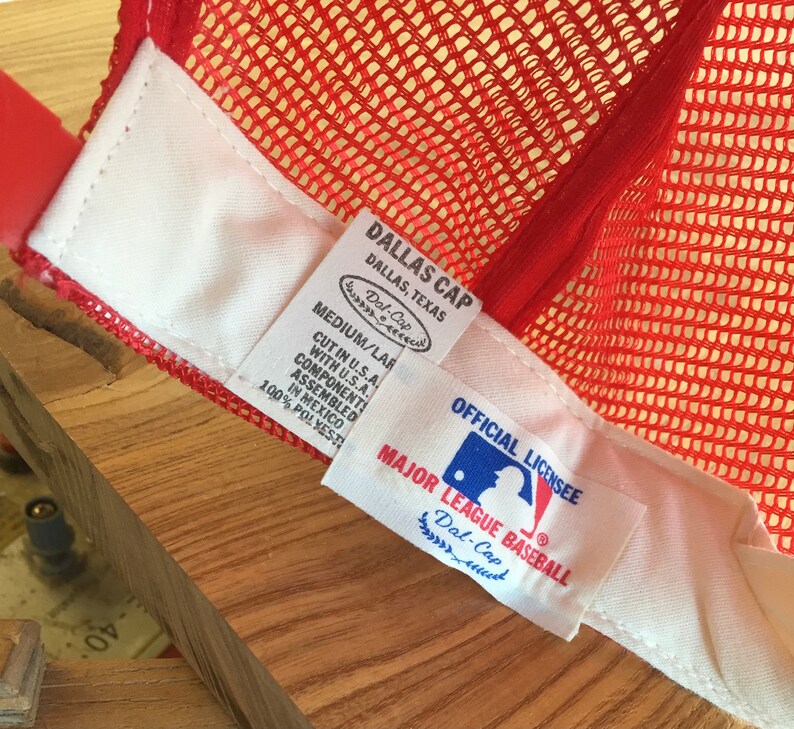 Puede incluir: Gorra de b&eacute;isbol de malla roja con una etiqueta blanca que dice "Dallas Cap, Dallas, Texas, Dal-Cap, Medium/Lar, Cut in U.S.A., Component with U.S.A., Assembled in Mexico, 100% Polyester" y una etiqueta blanca que dice "Official Licensee, Major League Baseball, Dal-Cap".