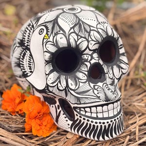 May include: A white ceramic skull sculpture featuring intricate black line art of flowers and a bird with a yellow beak. The skull has hollow eye sockets and a nasal cavity, with a painted toothy grin and decorative patterns covering the surface.
