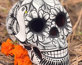 May include: A white ceramic skull sculpture featuring intricate black line art of flowers and a bird with a yellow beak. The skull has hollow eye sockets and a nasal cavity, with a painted toothy grin and decorative patterns covering the surface.
