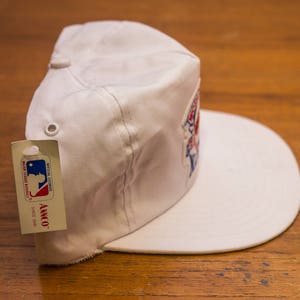 Pu&ograve; includere: Cappellino da baseball bianco con un logo rosso, bianco e blu e un'etichetta bianca che dice "Official Licensed Major League Baseball, Annco Since 1978".
