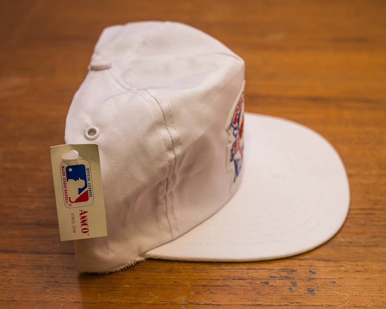 Pu&ograve; includere: Cappellino da baseball bianco con un logo rosso, bianco e blu e un'etichetta bianca che dice "Official Licensed Major League Baseball, Annco Since 1978".