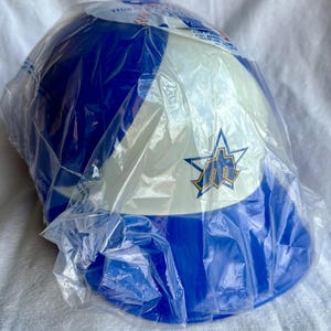 Op de afbeelding: Een blauw-witte baseballpet met een teamlogo, in een plastic verpakking. De pet heeft een blauwe kroon en een wit voorpaneel met een logo. De plastic verpakking is transparant.