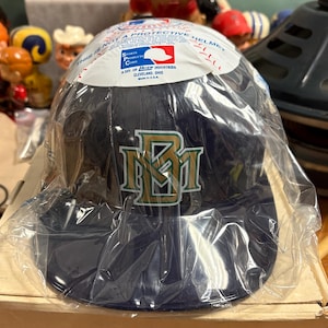 Op de afbeelding: Een marineblauwe baseballcap met goudgroene "NM" letters, nog in de originele verpakking. De cap heeft een gebogen klep en een beschermende sticker. De verpakking luidt: "Dit is geen beschermende helm."