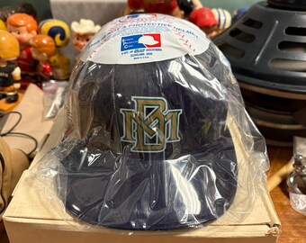 May include: A navy blue baseball cap with the letters "NM" in gold and green, still in its original packaging. The cap has a curved brim and a protective sticker on top. The packaging reads "This is not a protective helmet."