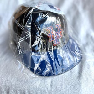 Op de afbeelding: Een zwarte baseballpet met een blauwe klep, met een oranje en blauw logo. De pet is verpakt in doorzichtig plastic. Het logo lijkt een gestileerd lettertype te zijn.