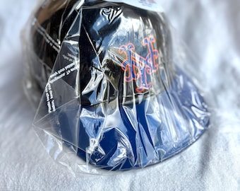 May include: A black baseball cap with a blue brim, featuring the New York Mets logo in orange and blue. The cap is wrapped in clear plastic packaging.