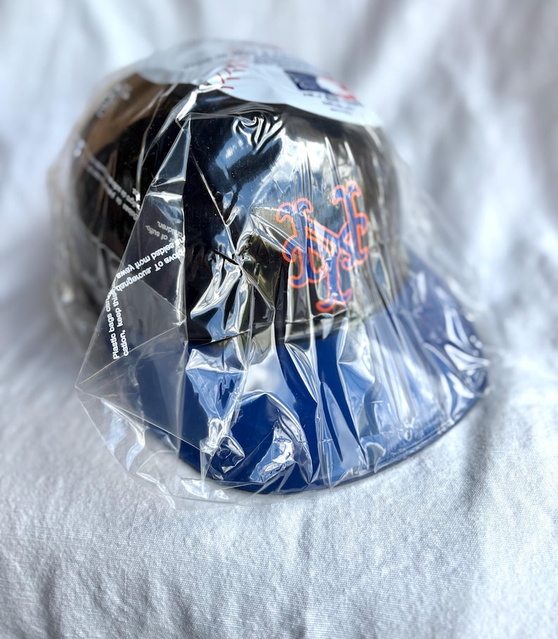 Op de afbeelding: Een zwarte baseballpet met een blauwe klep, met een oranje en blauw logo. De pet is verpakt in doorzichtig plastic. Het logo lijkt een gestileerd lettertype te zijn.
