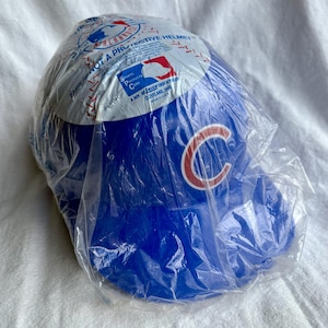 Op de afbeelding: Een blauwe honkbalhelm, verpakt in doorzichtig plastic. De helm heeft een rood en wit "C"-logo en de tekst "THIS IS NOT A PROTECTIVE HELMET". De verpakking toont ook het MLB-logo.