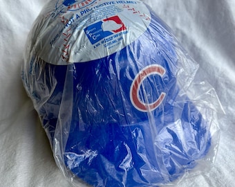 May include: A blue baseball helmet, packaged in clear plastic. The helmet features a red and white "C" logo and text that reads "THIS IS NOT A PROTECTIVE HELMET". The packaging also displays the MLB logo.
