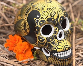 May include: A black skull sculpture decorated with intricate gold painted floral patterns and a bird motif. The eye sockets and nasal cavity are outlined in white, and the teeth are painted in white. Two bright orange marigold flowers are placed nearby.