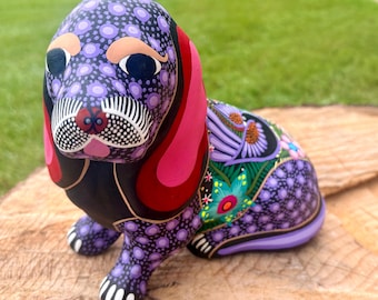 May include: A hand-painted wooden dog figurine featuring a purple and black spotted pattern, long bright red ears, and intricate floral designs in shades of teal, pink, and orange on its back. The dog has white painted claws and black eyes.
