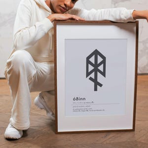 May include: A framed print featuring the word "óðinn" and a stylized rune symbol in dark gray. The print has a white background and a brown wooden frame. The text below the symbol describes the meaning of the word.
