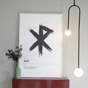 Könnte beinhalten: Ein weiß gerahmter Kunstdruck mit einem großen schwarzen Symbol und dem Wort "ást" unten. Der Text darunter lautet "love" und beschreibt die Runen Gebo und Wunjo. Eine grüne Vase mit Grün steht auf einer roten Oberfläche.