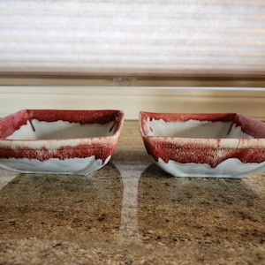 May include: Two square ceramic bowls with a white glaze and red accents. The bowls are slightly angled and have a rustic, handmade look.