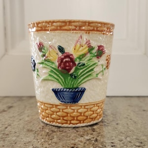 May include: A white ceramic cup with a floral design. The cup has a basket weave pattern around the top and bottom. The flowers are in a blue and white pot.