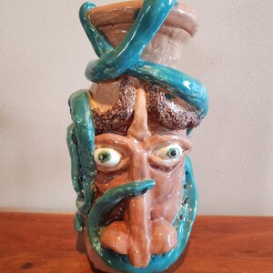 May include: A ceramic vase with a face design, featuring a brown face with green eyes and a long nose. Turquoise snakes wrap around the vase. The top of the vase is a light brown color.