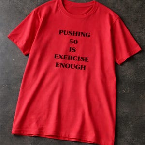 May include: A bright red t-shirt with the text "PUSHING 50 IS EXERCISE ENOUGH" printed in black. The short-sleeved shirt is made of a soft material and is displayed on a dark gray surface.