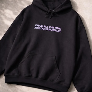 May include: Black hooded sweatshirt with a front pocket and drawstring. The text "DISCO ALL THE TIME. KISS, OCCASIONALLY." is printed in a light purple color on the front. The sweatshirt is made of a soft material.