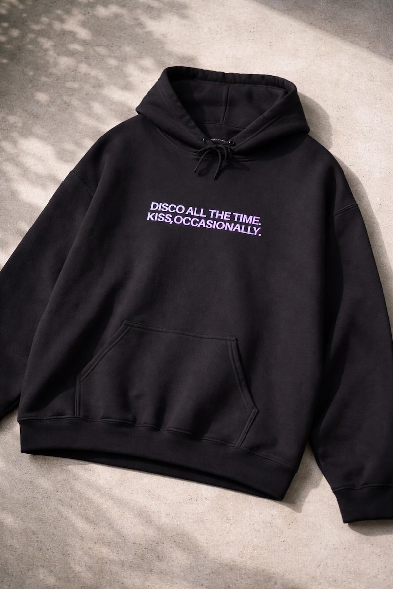 May include: Black hooded sweatshirt with a front pocket and drawstring. The text "DISCO ALL THE TIME. KISS, OCCASIONALLY." is printed in a light purple color on the front. The sweatshirt is made of a soft material.