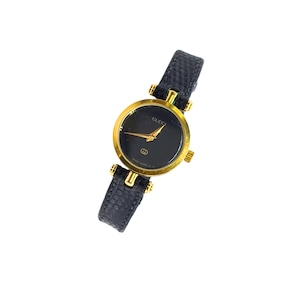 Puede incluir: Un reloj de pulsera Gucci con caja redonda en tono dorado y esfera negra con manecillas doradas. El reloj tiene una correa de cuero negro texturizado con herrajes dorados que conectan la correa a la caja del reloj en ambos lados.