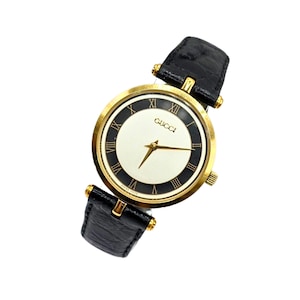 Pode incluir: Um relógio de pulso Gucci com caixa redonda dourada, mostrador branco com anel externo preto e numerais romanos. O relógio inclui ponteiros dourados e uma pulseira de couro preto texturizado com ferragens em metal dourado.