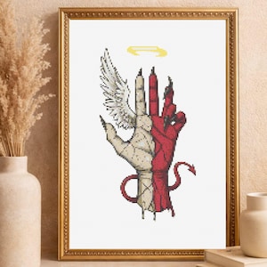 Puede incluir: Obra de arte en punto de cruz enmarcada con un diseño de mano dividida. Un lado es blanco con un ala, el otro rojo con una cola de diablo. Un halo amarillo está encima de la mano. La obra está en un marco dorado.