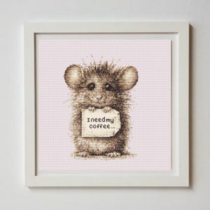 May include: A framed cross-stitch artwork featuring a brown mouse holding a sign that reads "I need my coffee...". The mouse has large ears and a fluffy appearance. The background is a light pink color.